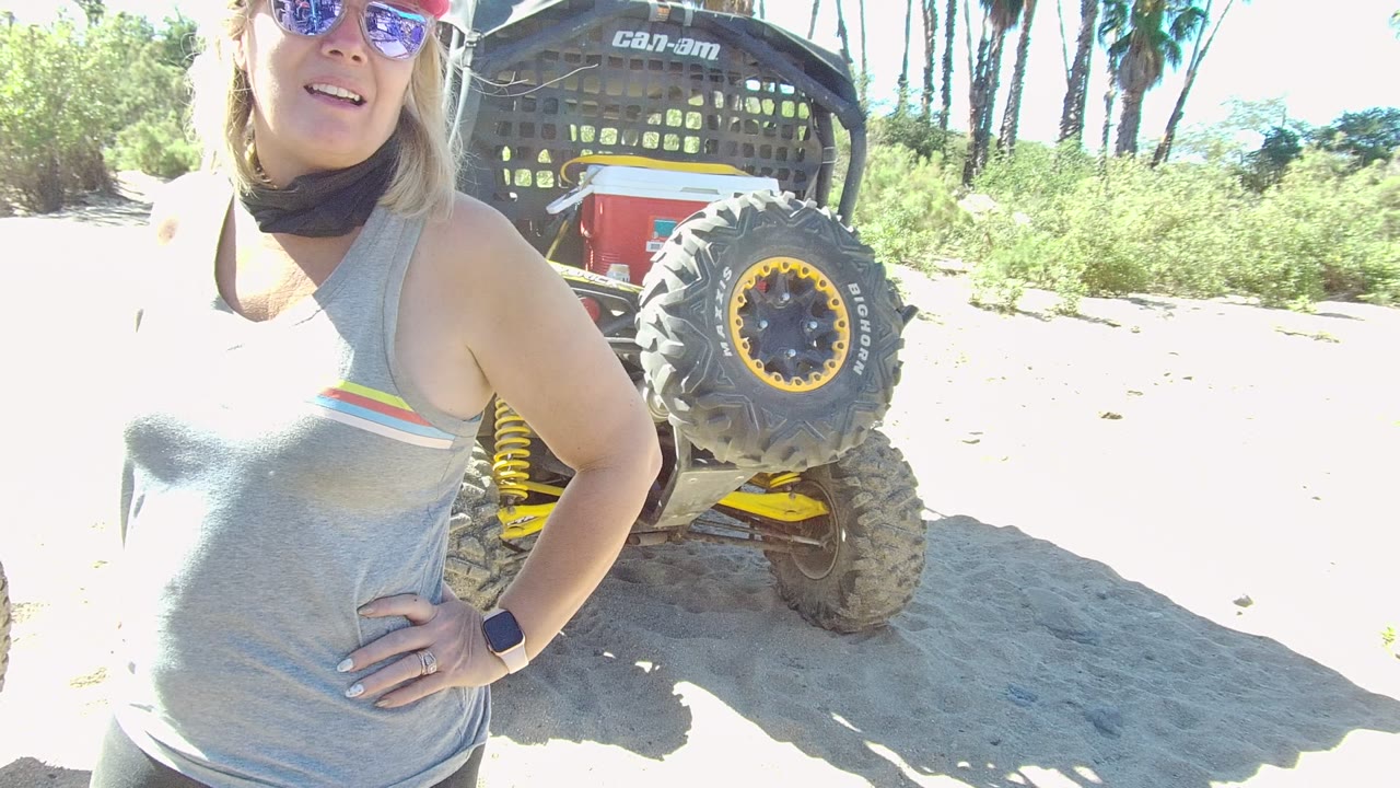 In Los Cabos, a woman in sunglasses and a gray tank top stands close to camera with a Can-Am off-road vehicle wheel behind her on bright sandy ground.