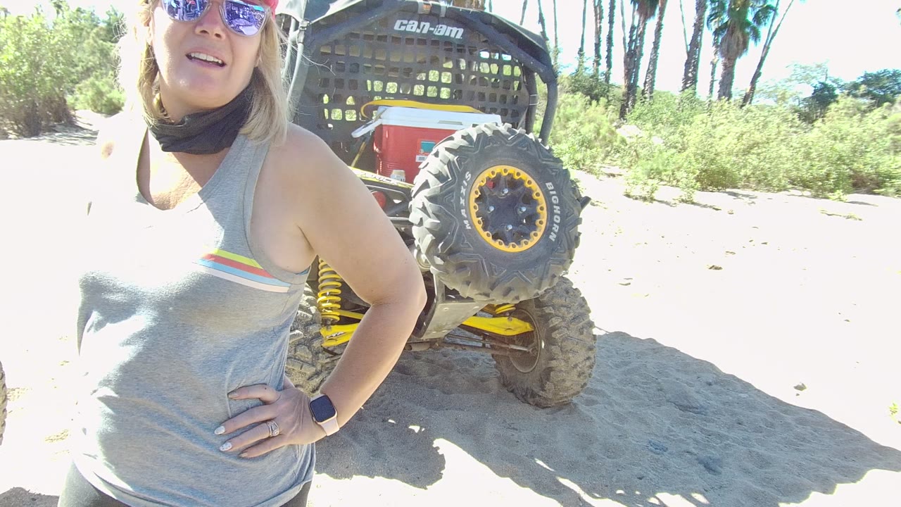 In Los Cabos, the woman faces slightly toward camera beside the black roll cage and yellow-accented wheel of the off-road vehicle, with palms and green vegetation behind.