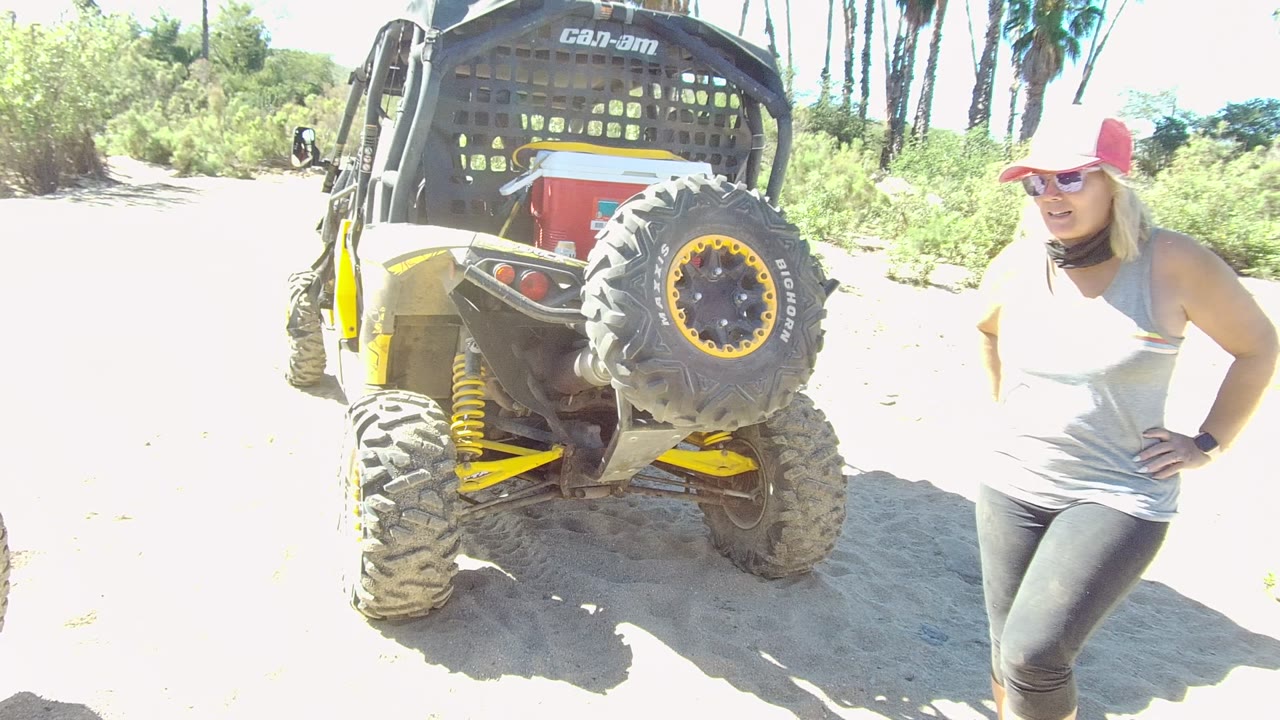 In Los Cabos, Baja California Sur, Mexico, a woman in a red cap and sunglasses stands near the rear tire of a Can-Am off-road vehicle on bright sandy ground.