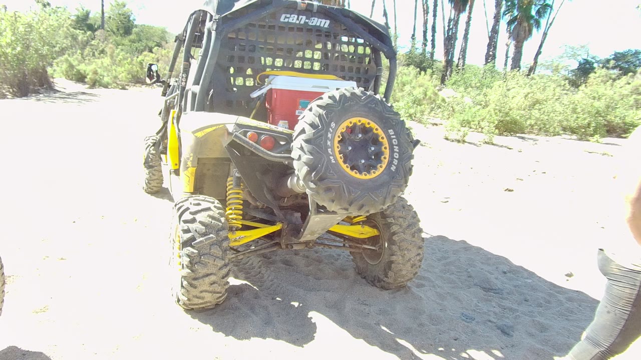The parked UTV remains centered on the sandy Los Cabos path, with its spare tire, yellow suspension, red cooler, bright ground, and palms behind it.