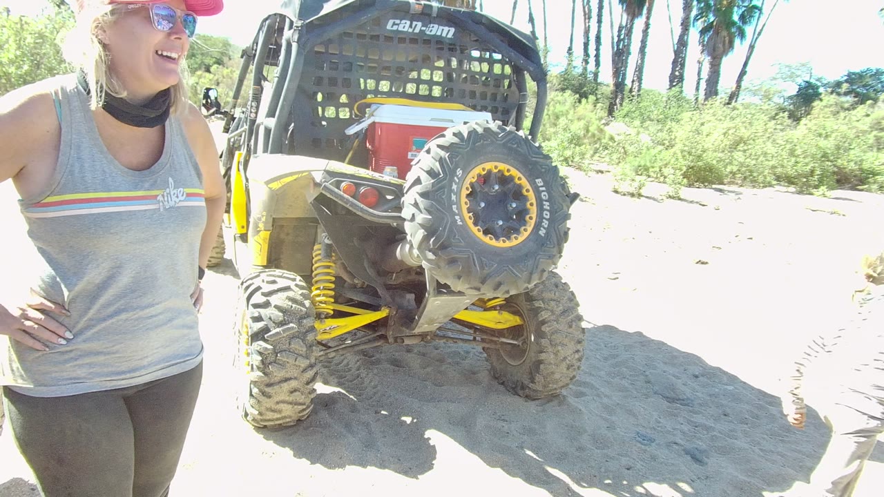 In Los Cabos, Baja California Sur, a woman in a red cap and sunglasses stands near the rear tire of a parked dune buggy on bright sandy ground.