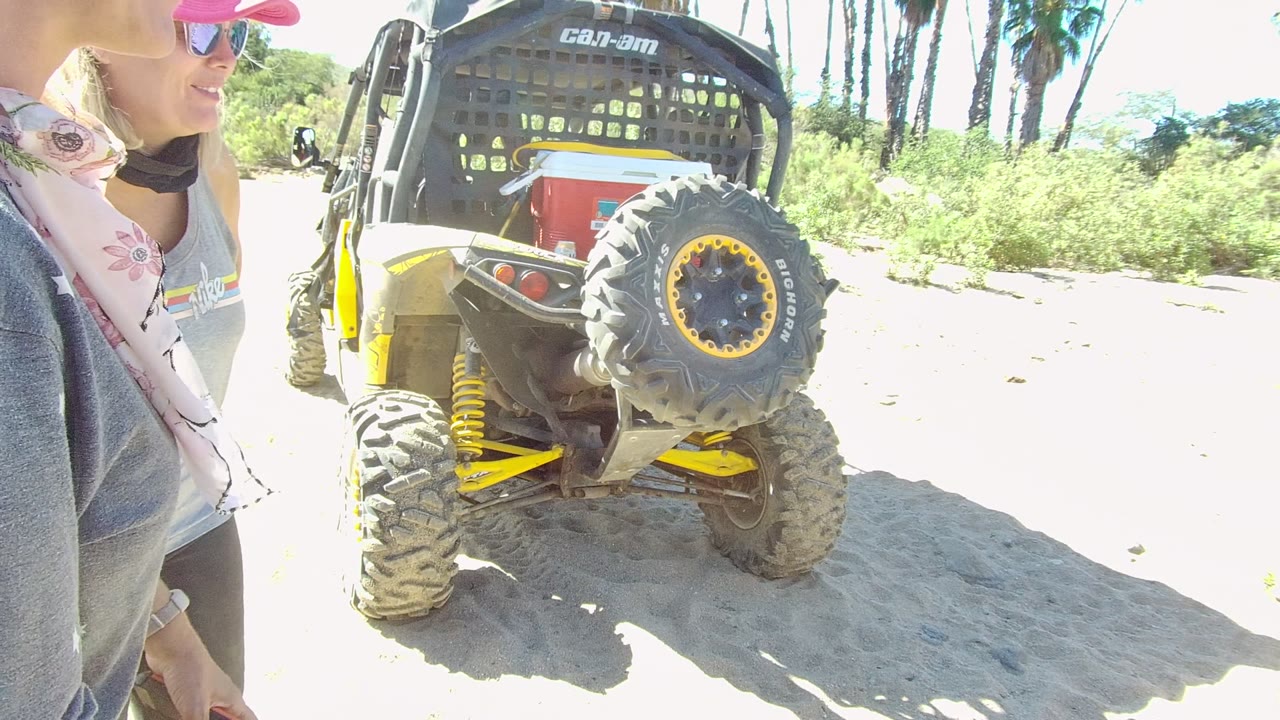 Two adults near the rear of a parked Can-Am dune buggy on sand.