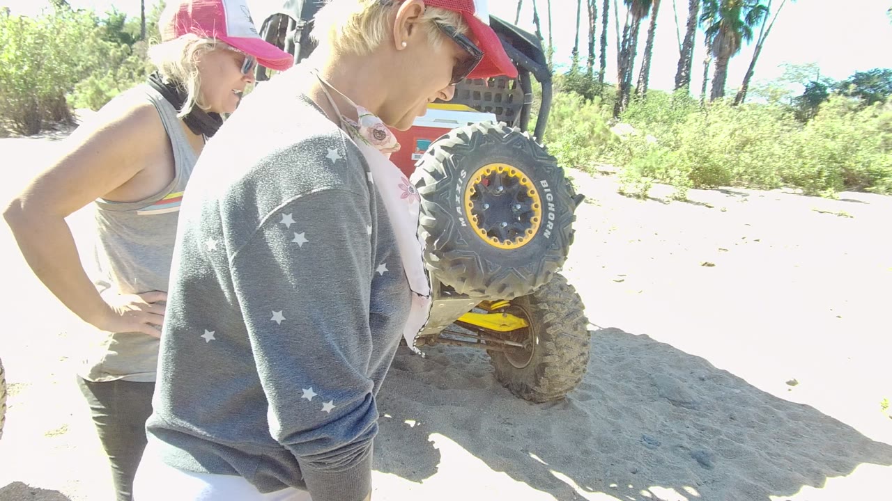 In Los Cabos, Baja California Sur, the woman in a red cap and another adult stand behind the buggy as sandy terrain, green vegetation, and tall palms remain in view.