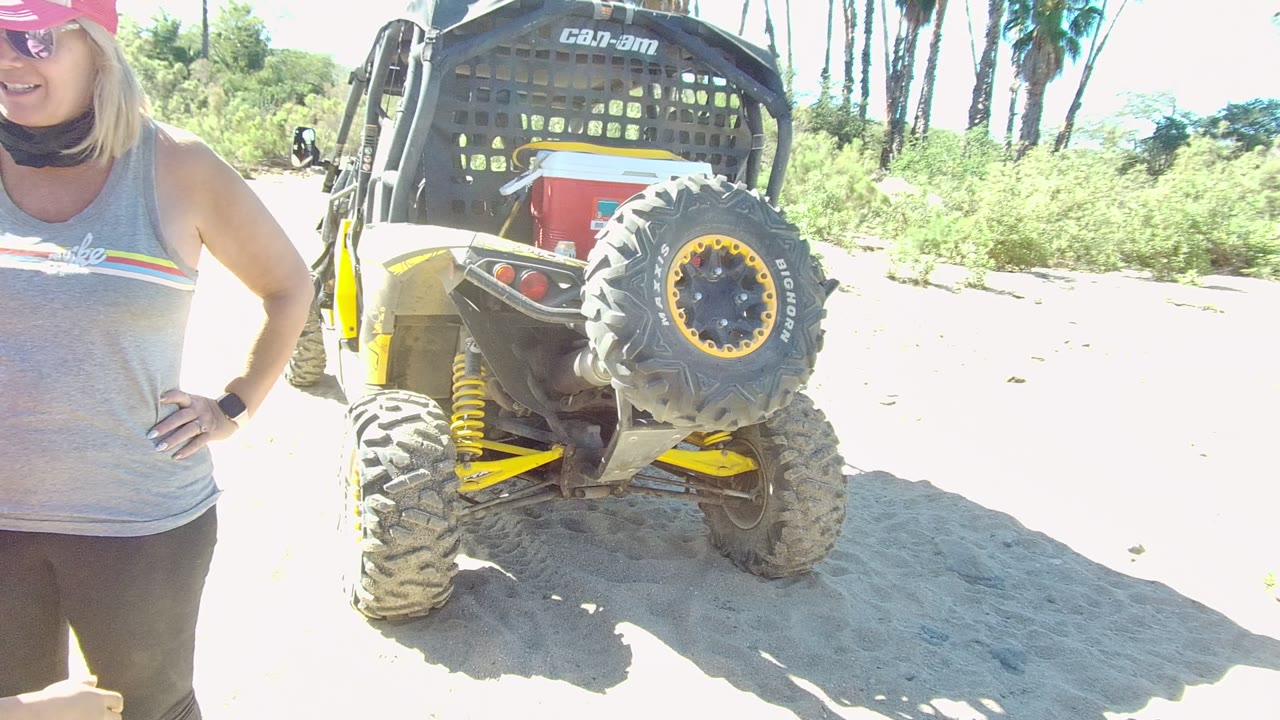 In Los Cabos, a woman in a gray tank top and pink cap stands beside the rear of a parked off-road vehicle on bright sand, with vegetation behind it.