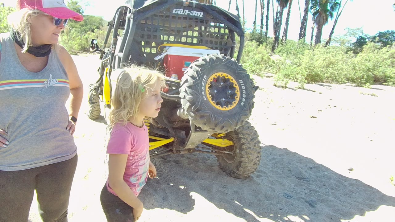 The woman looks toward a child in a pink shirt beside the off-road vehicle in Los Cabos, with the vehicle's spare tire and roll cage prominent in the frame.