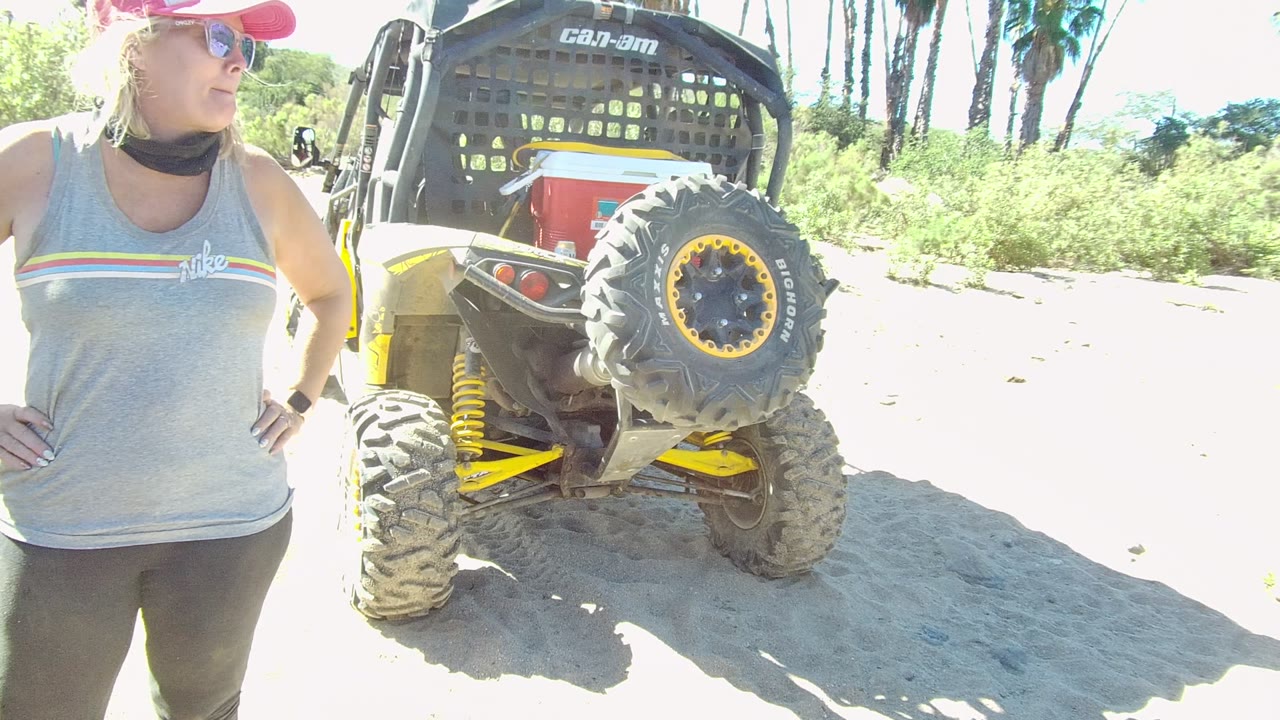 At 0:02, the woman looks off to the side while standing next to the rear of the off-road vehicle, with overexposed sand and palm trunks in the Los Cabos background.