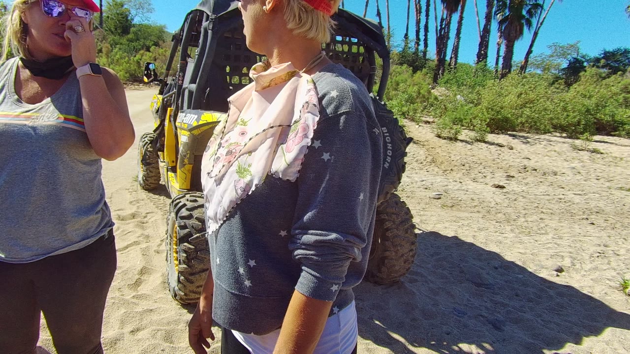 Two women continue talking near a yellow dune buggy on sandy Los Cabos terrain, with one hand raised near a face covering and palms behind them.