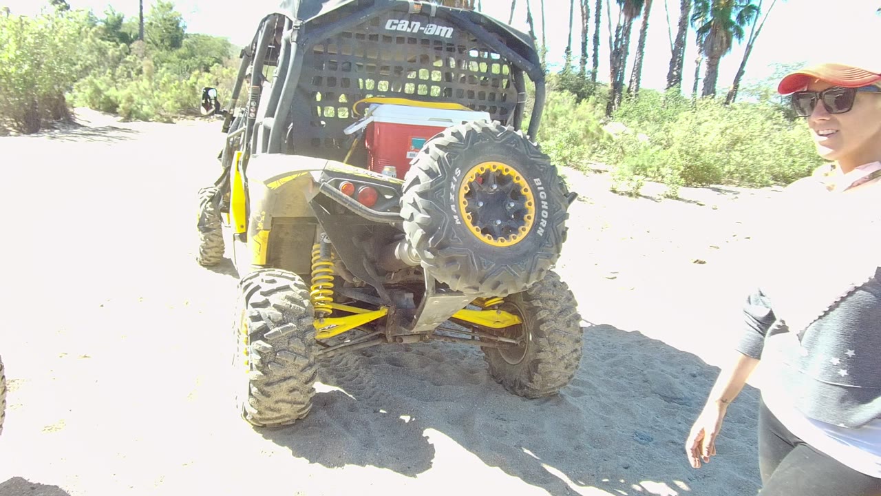 A woman wearing a red cap and sunglasses appears at the right edge beside the parked off-road vehicle in Los Cabos, with shrubs and palms behind the sandy path.