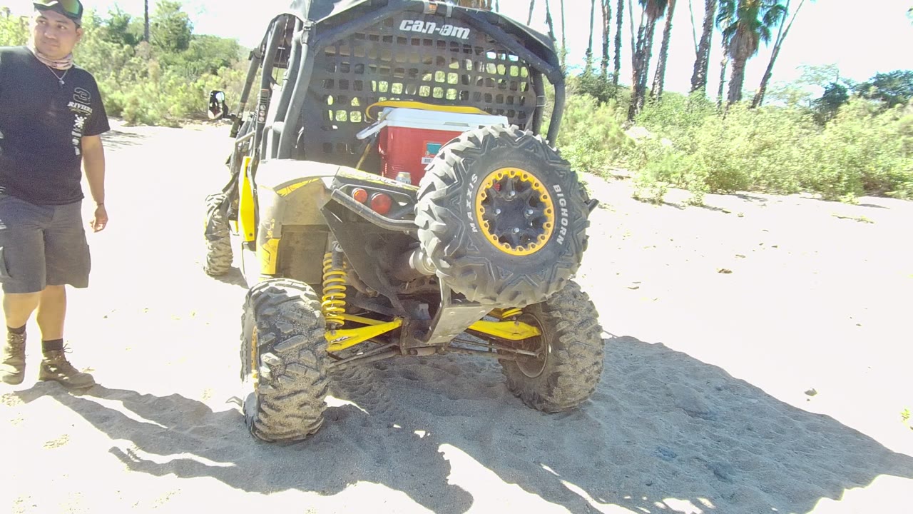 A man in a black shirt, gray shorts, boots, and cap stands beside the parked Can-Am off-road vehicle on sandy ground in Los Cabos, Mexico.