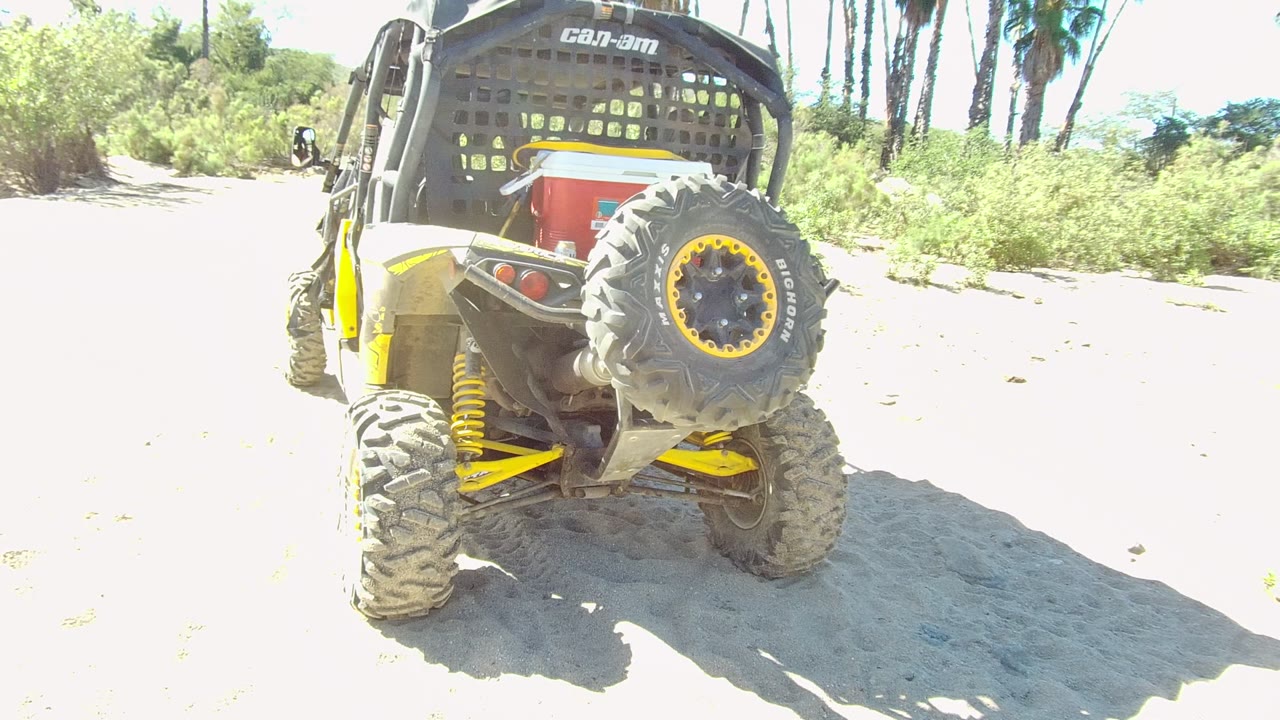 At 0:00, a rear view of a Can-Am UTV with yellow suspension and a spare tire sits on a sandy dirt path in Los Cabos, Mexico, with no visible people.