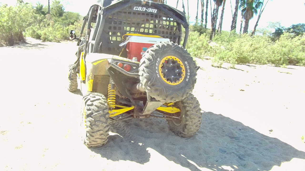 At 0:02, the black and yellow Can-Am UTV is seen from the rear on a sunlit sandy path in Los Cabos, backed by shrubs and palms, with no visible people.