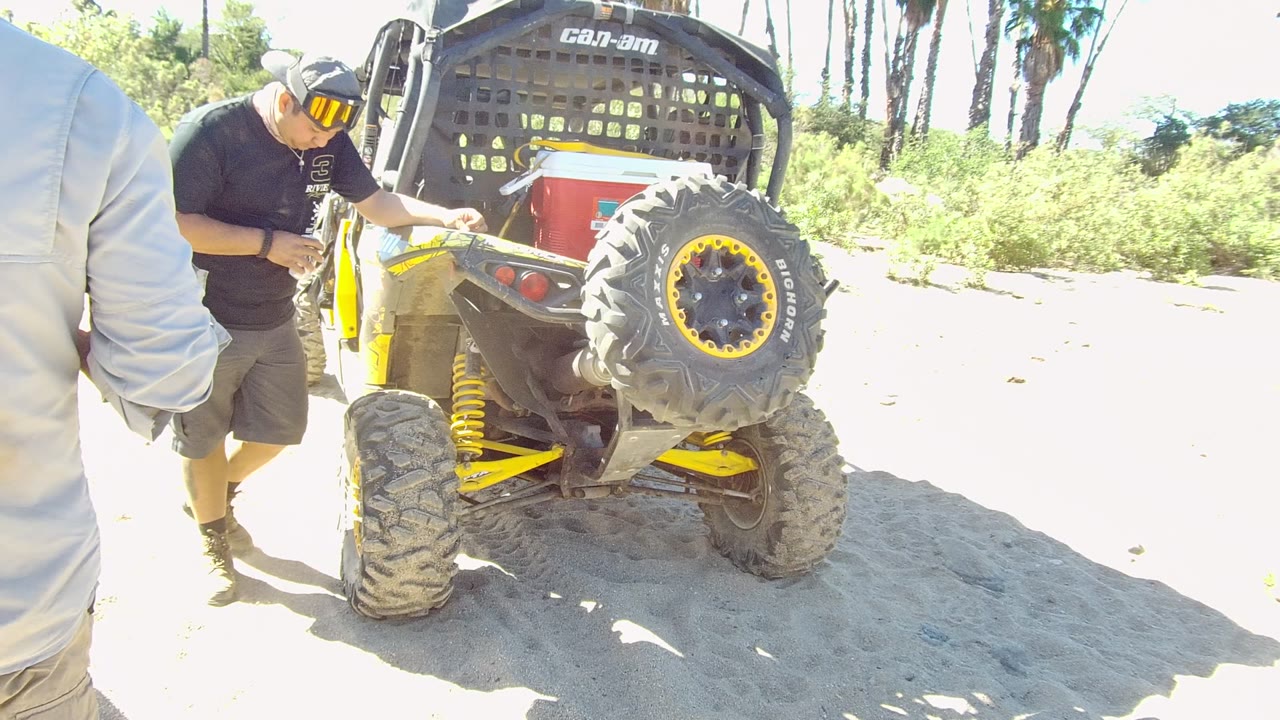 At 0:00 in Los Cabos, Baja California Sur, a man stands beside the rear of a yellow and black UTV on bright sandy ground with vegetation behind it.