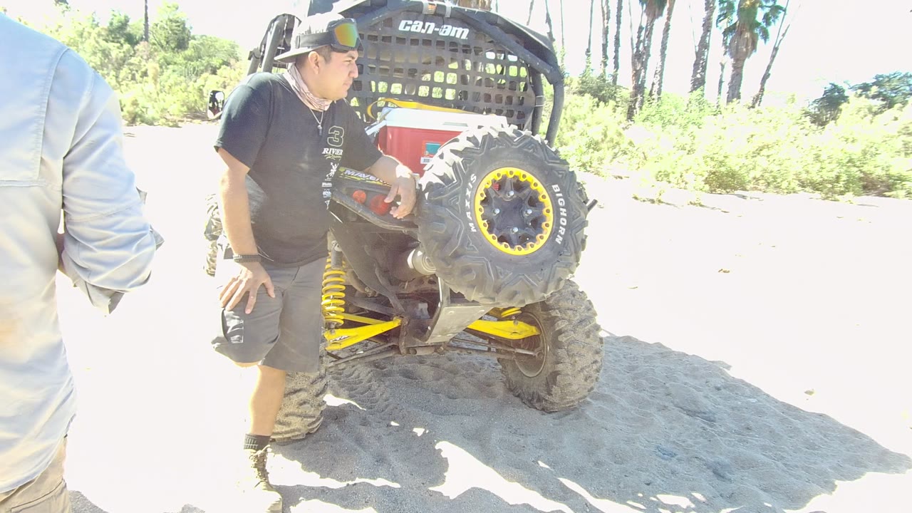 In Los Cabos, Baja California Sur, Mexico, a man in a black shirt and cap stands beside the rear of a small off-road vehicle on pale sand with bright greenery behind him.