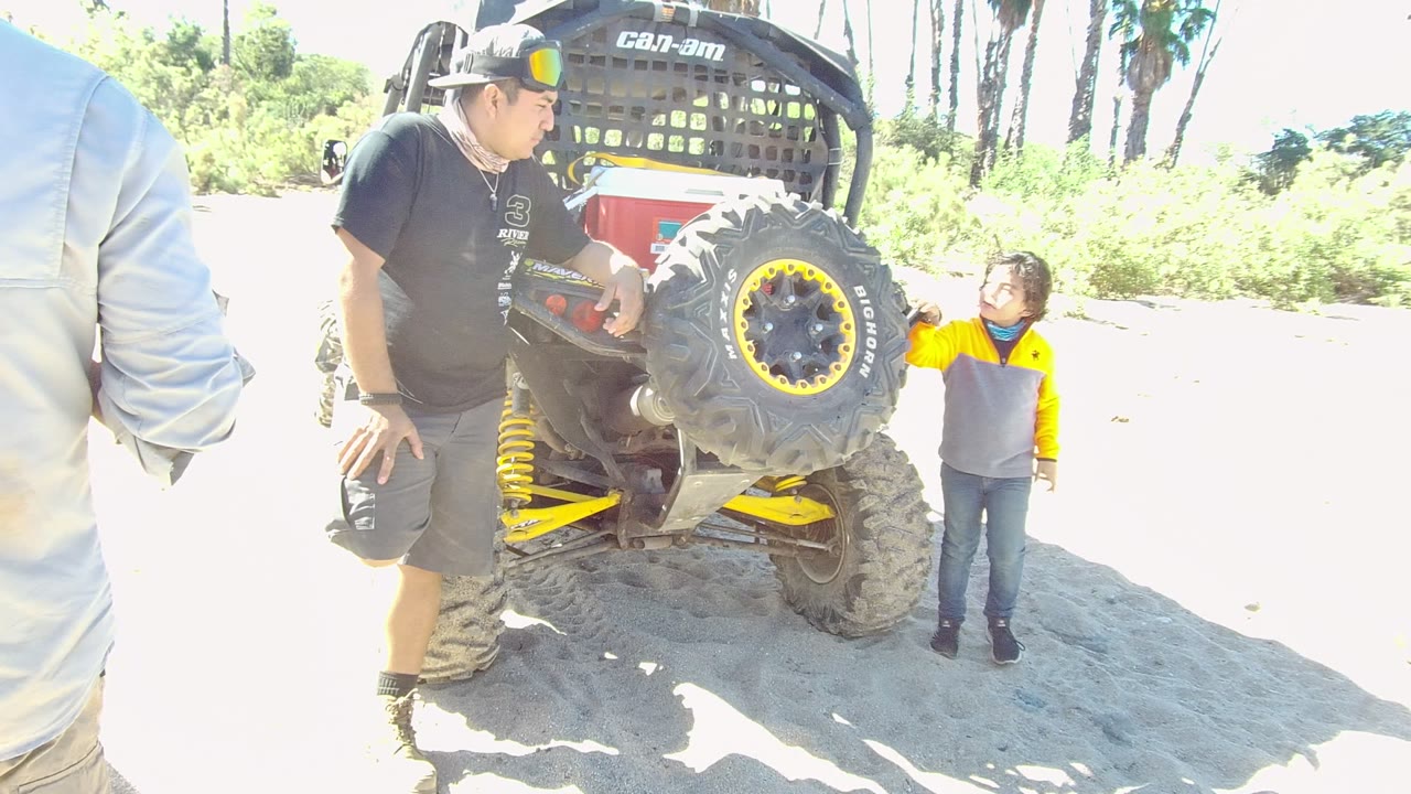 In Los Cabos, Baja California Sur, Mexico, the child stands close to the rear tire and reaches toward the small off-road vehicle while the man watches from the side.