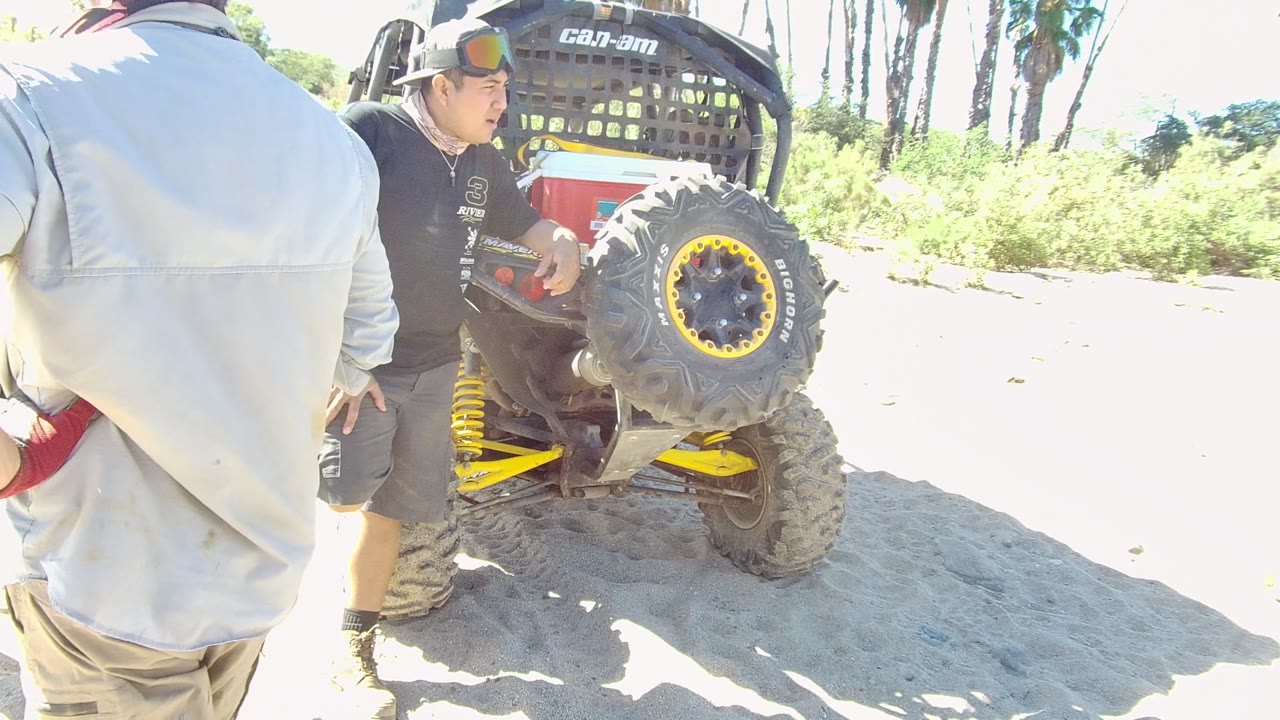 In Los Cabos, a man in a black shirt and cap stands near the rear tire of a red and yellow utility vehicle while a light-jacketed person is cropped in the foreground.