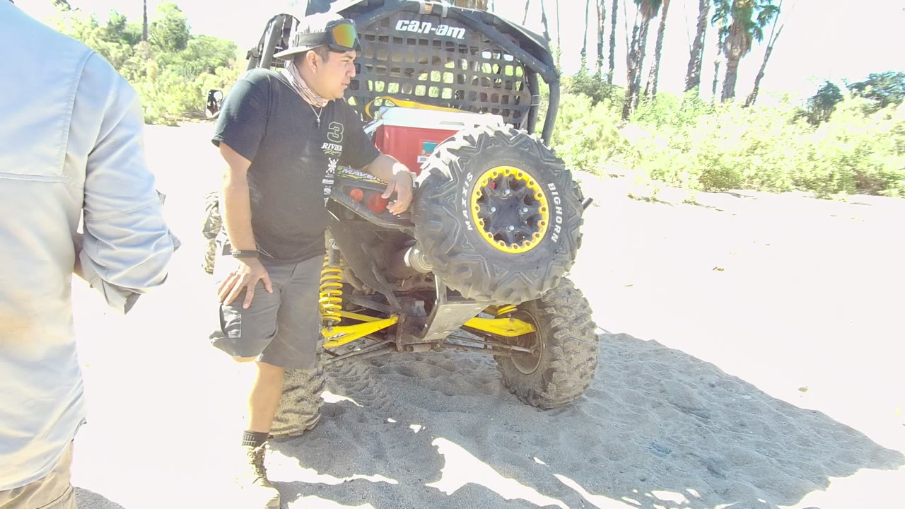 In Los Cabos, a cropped person in a light jacket stands at left while the man in black stays near the rear tire of the utility vehicle under harsh bright light.