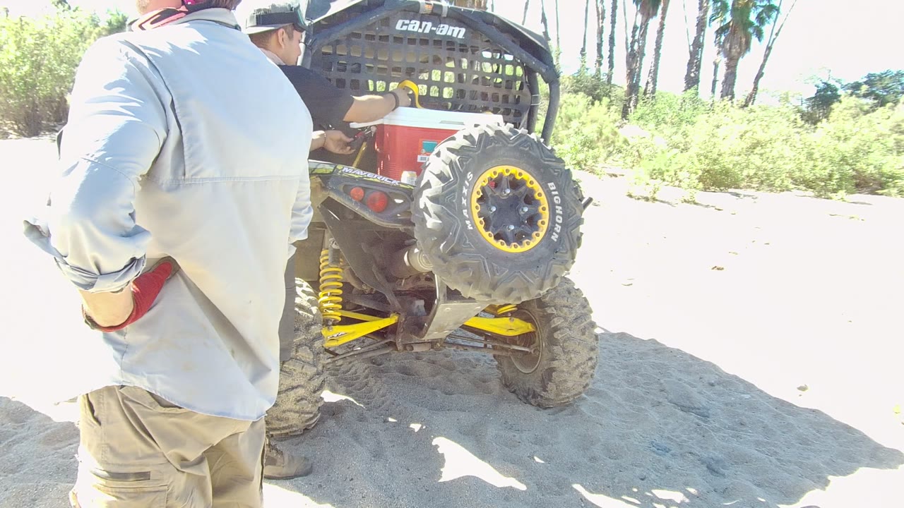 At 0:02 in Los Cabos, the rear of the red and yellow off-road vehicle stays centered, with one man partly blocking the left side and brush visible beyond the sand.