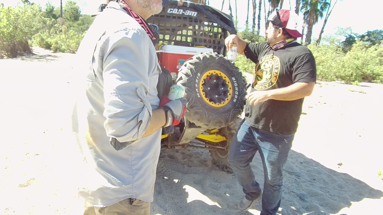 In Los Cabos, Baja California Sur, two adult men shake hands beside the rear tire of a Can-Am off-road vehicle on bright sandy ground.