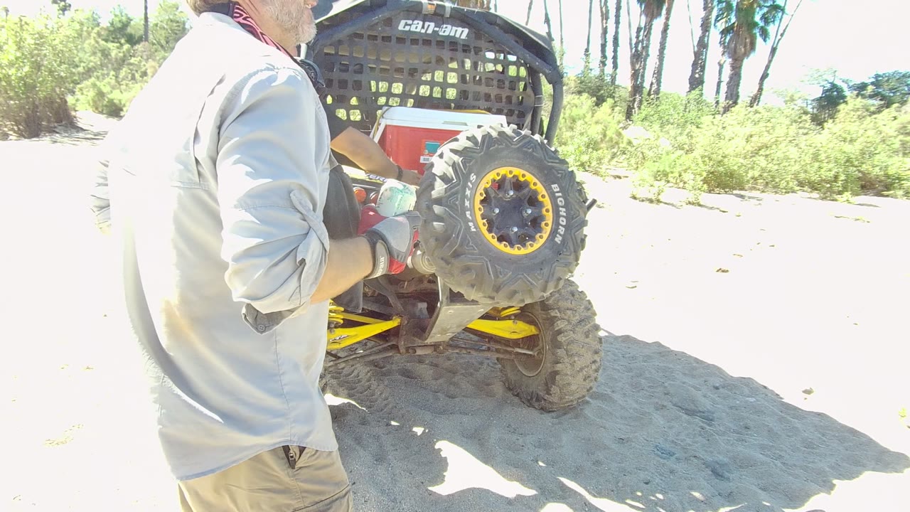 At 0:02 in Los Cabos, Baja California Sur, the foreground man remains beside the ATV spare tire, with bright sun washing the sand and vegetation in the background.