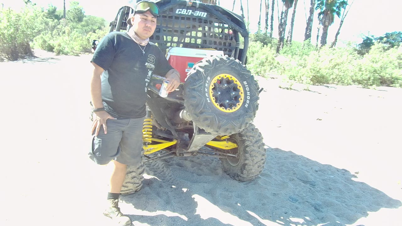 In Los Cabos, a man in dark casual clothing stands beside the rear tire and spare wheel of an off-road vehicle on bright sandy ground.