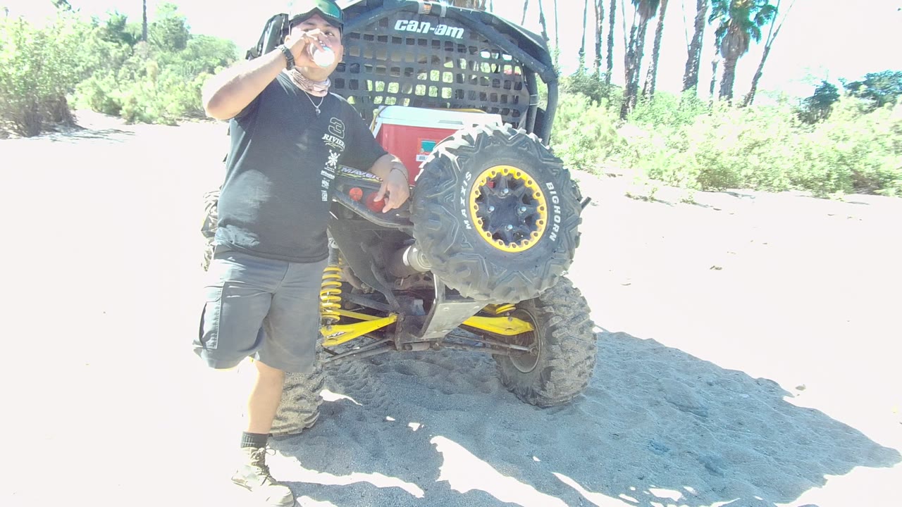 At 0:00 in Los Cabos, a man raises a bottle to his mouth beside the rear of a Can-Am off-road vehicle on bright sandy ground.