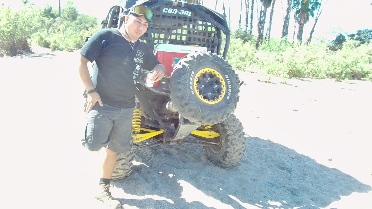 At 0:02 in Los Cabos, the man stands closer to the ATV with the bottle near his chest, framed against washed-out sand and background palms.