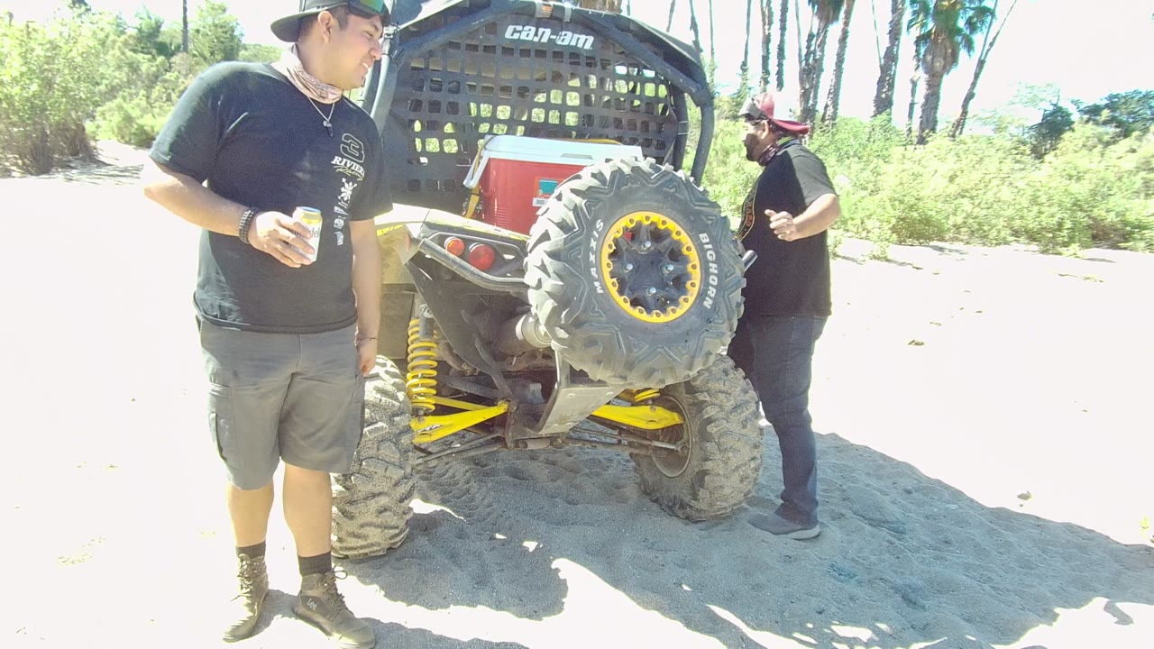 In Los Cabos, Baja California Sur, two men stand near the rear of an off-road vehicle, with the main man holding a cup and the spare tire centered beside bright sandy ground.