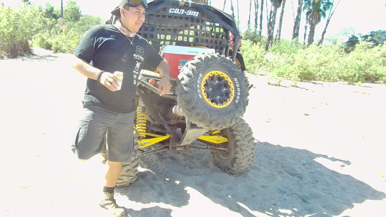 In Los Cabos, Baja California Sur, the man shifts his stance beside the off-road vehicle, holding a cup with one leg raised near the sandy ground and bright vegetation behind him.