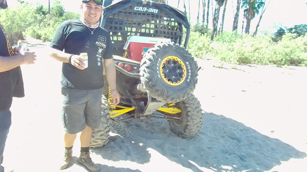 In Los Cabos, a man in a dark shirt and shorts stands beside the rear tire and spare tire of a dune buggy on bright sandy ground.