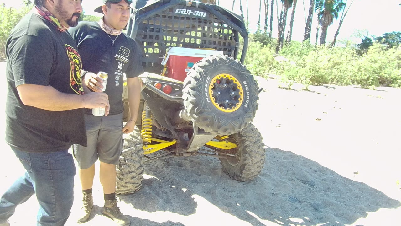 In Los Cabos, two adult men stand beside the rear of a dune buggy, with the branded roll cage, spare tire, and yellow suspension visible in bright light.