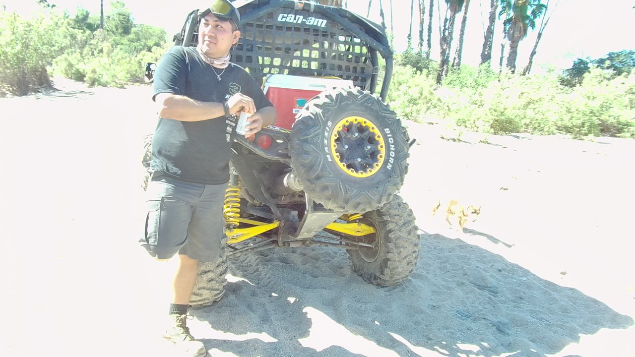 A man stands next to a sandy ATV in Los Cabos, Baja California Sur, with bright sunlight washing the ground and green vegetation behind him.