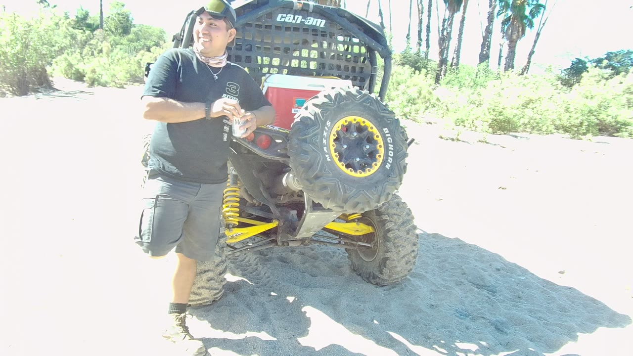 At 0:00 in Los Cabos, a man in a black shirt and cap stands beside a red and black ATV with a large yellow-rimmed tire on bright sand.