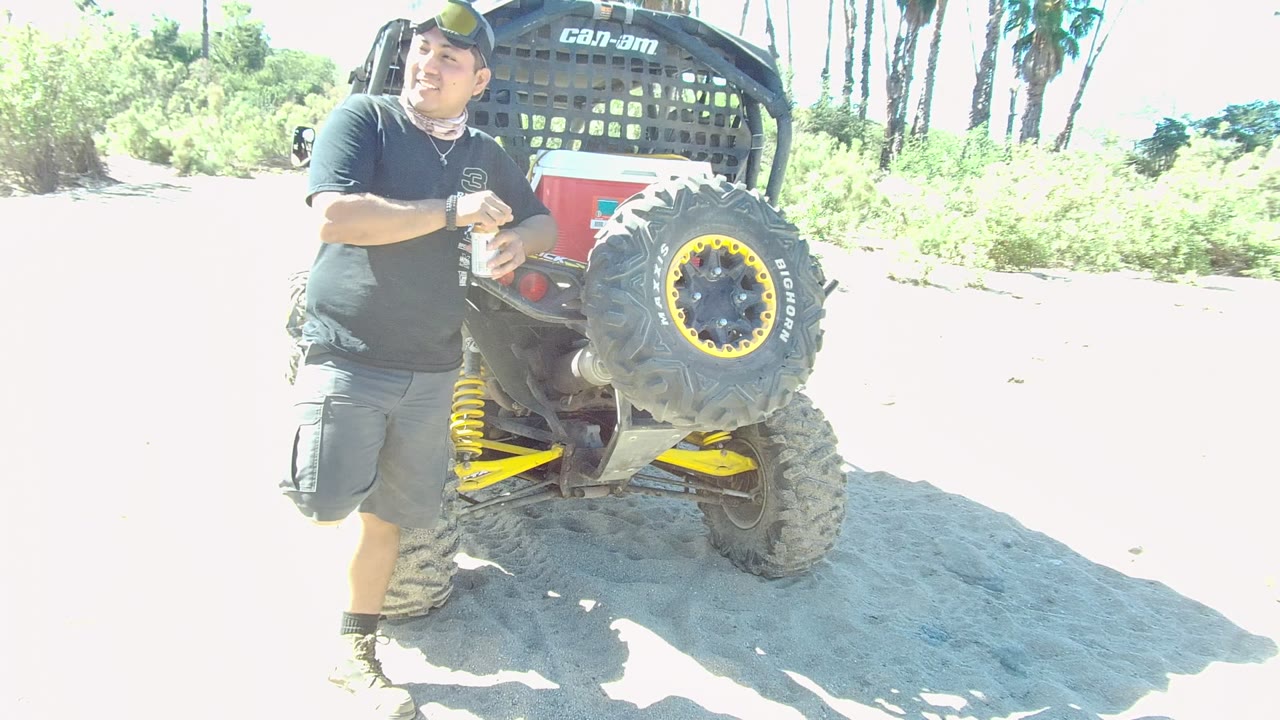 At 0:02 in Los Cabos, the man looks off to the side with one hand near the ATV frame, while the vehicle tire, yellow suspension, sand, shrubs, and palms remain visible.