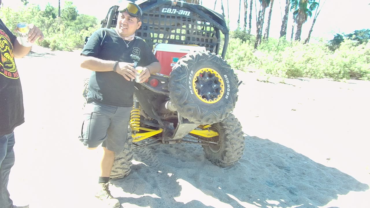 At 0:00 in Los Cabos, a man in a black shirt holds a drink near the rear of a Can-Am off-road vehicle while another person is partly cropped at the left edge.