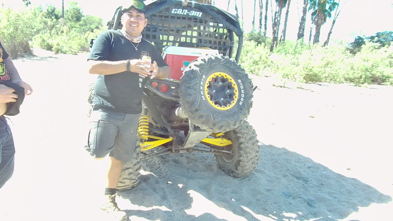 At 0:02 in Los Cabos, a person in a black shirt is cropped at the left while the sandy track and greenery dominate the frame, with only part of the off-road vehicle still visible.