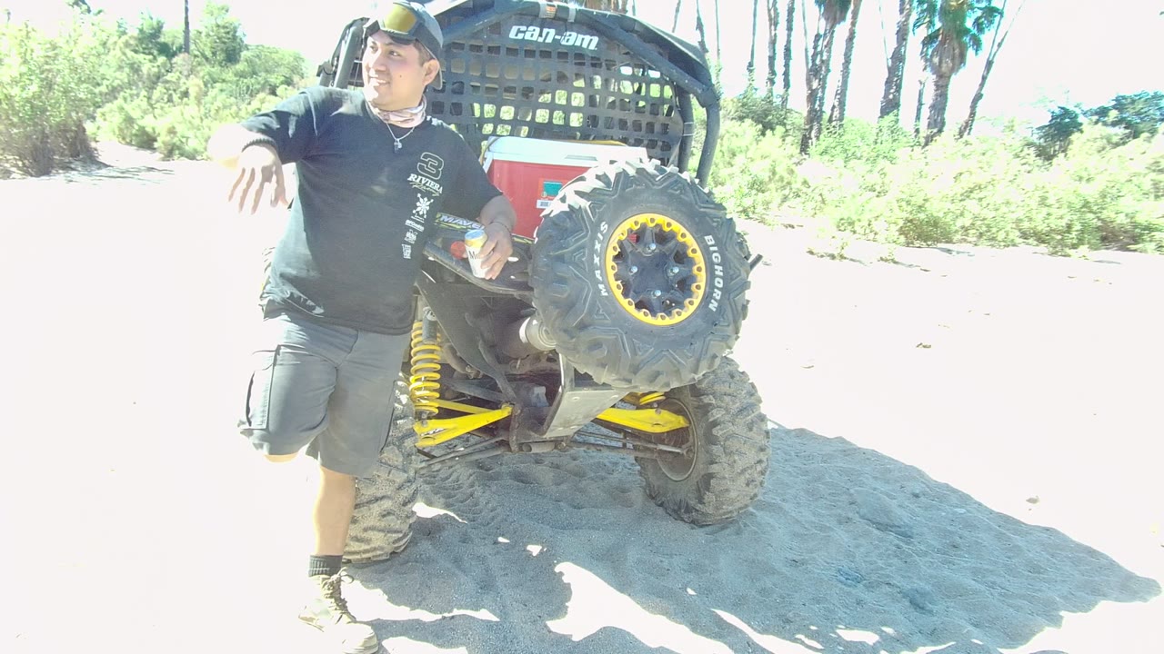 In Los Cabos, a man in dark casual clothing stands beside the rear of a dusty off-road vehicle on a bright sandy roadside with greenery behind him.