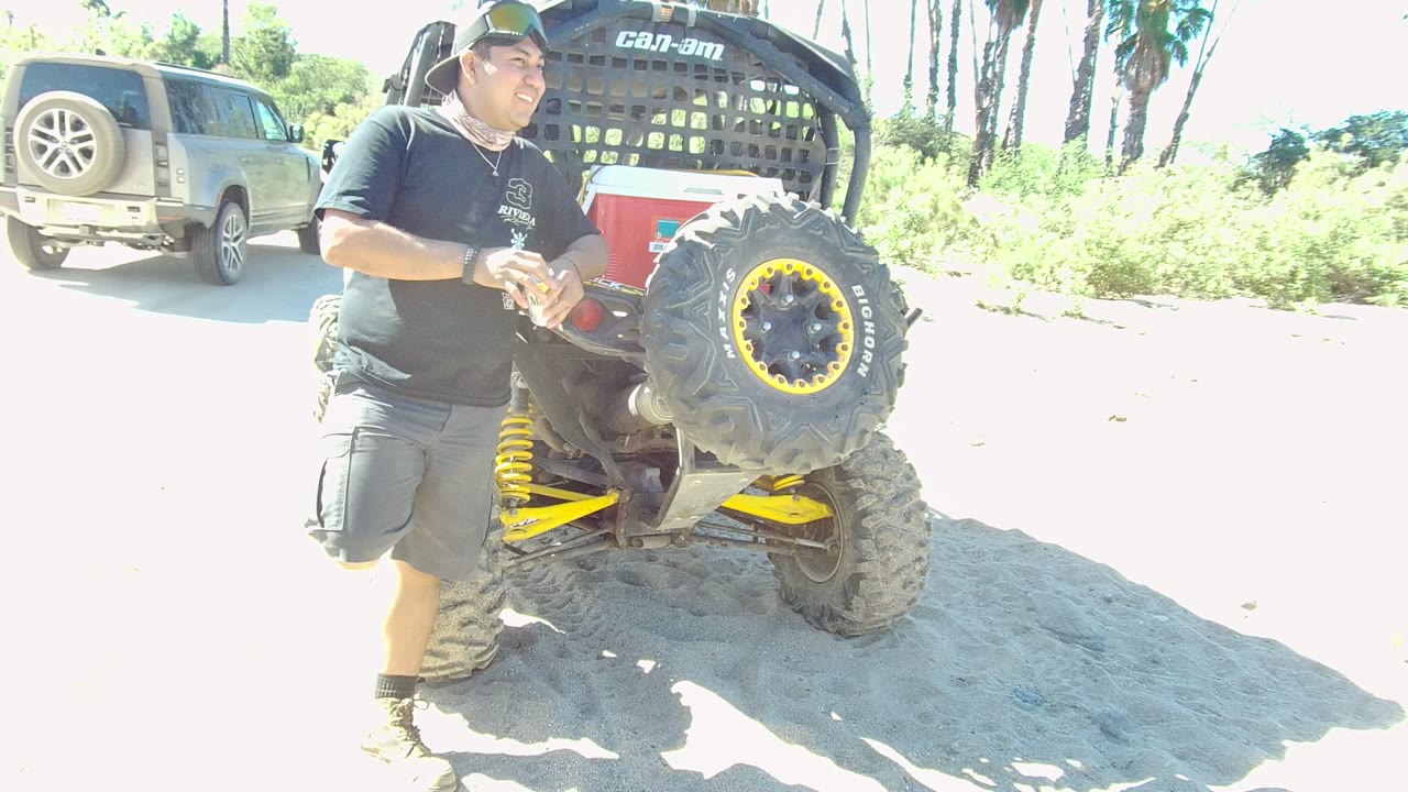In Los Cabos, a gray vehicle passes in the background while the man stands beside the off-road vehicle in strong sun on a sandy roadside.