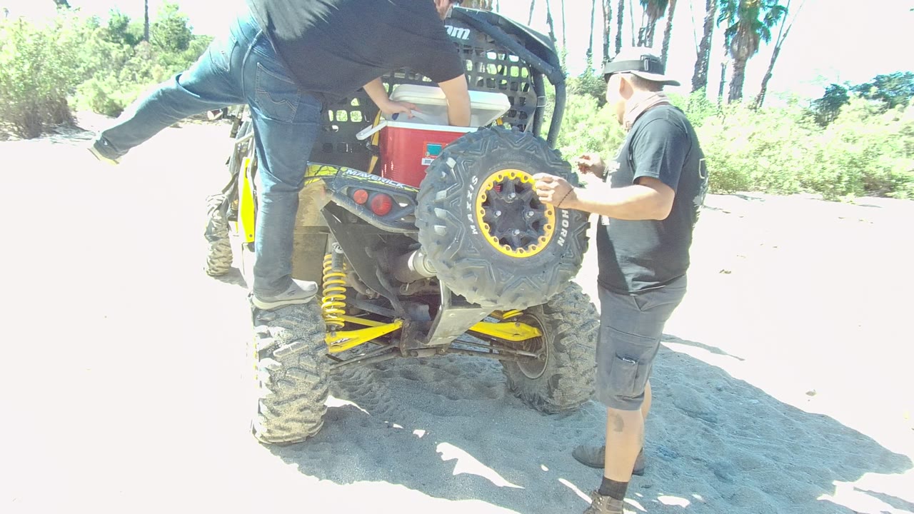 At 0:00 in Los Cabos, a man begins stepping down from the rear of a yellow and black ATV while another man stands by the spare tire on sandy ground.