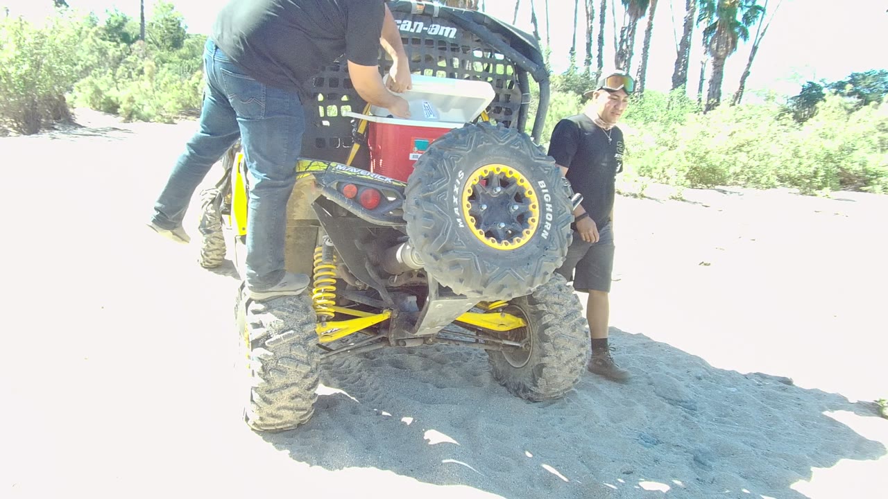 At 0:00 in Los Cabos, Baja California Sur, one man stands on the rear of a Can-Am ATV while another stands nearby on bright sandy ground with greenery behind.