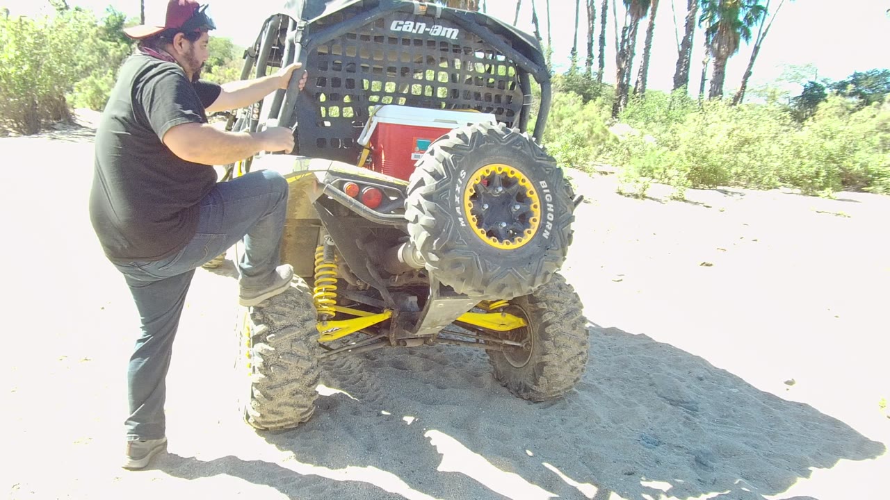 In Los Cabos, Baja California Sur, a person in a black shirt and cap steps onto the rear tire of a small off-road vehicle on bright sandy ground.