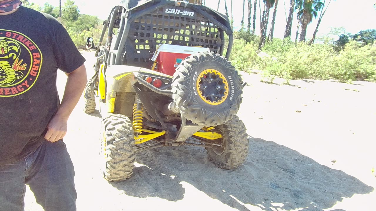 In Los Cabos, a cropped man stands at the left edge beside the rear of a Can-Am off-road vehicle on bright sandy ground with greenery and palms behind it.