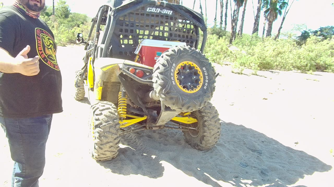 In Los Cabos, the man gives a thumbs-up while standing beside the rear of the Can-Am UTV, with the spare tire, yellow suspension, sand, greenery, and palms visible.