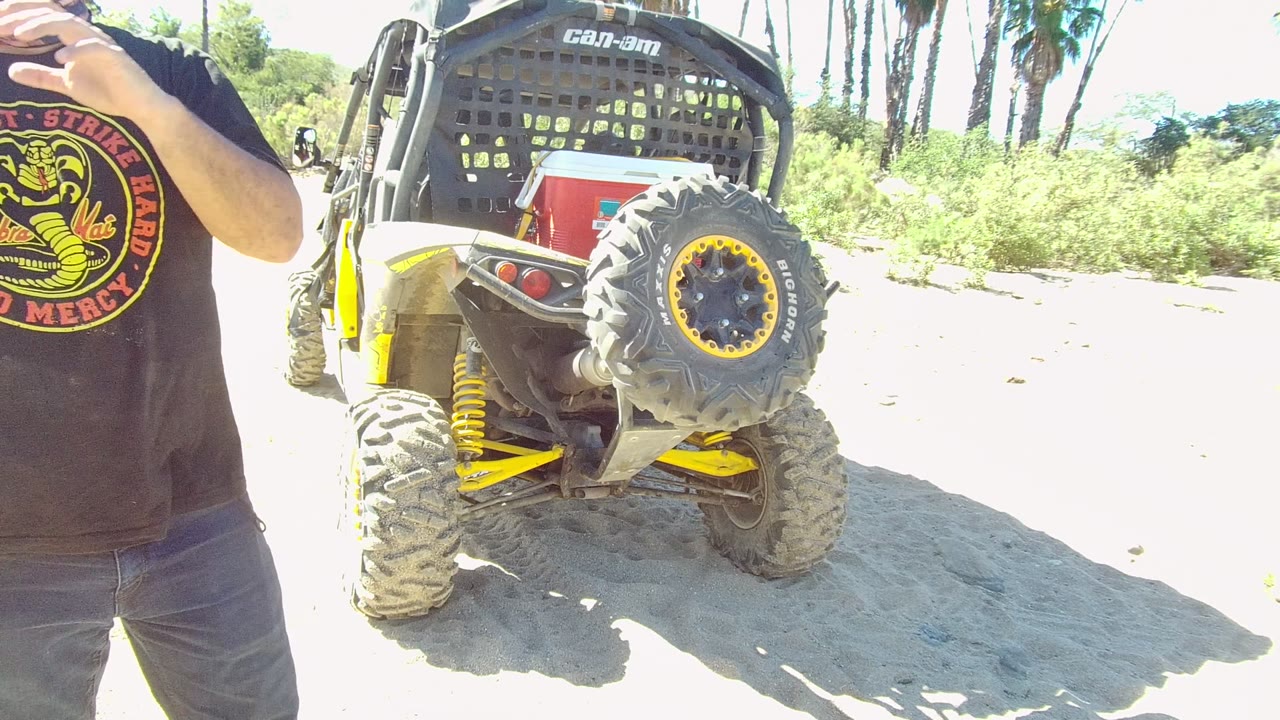 The Los Cabos frame shows the Can-Am UTV rear and spare tire centered while the cropped person in a black logo shirt gestures near the right edge.