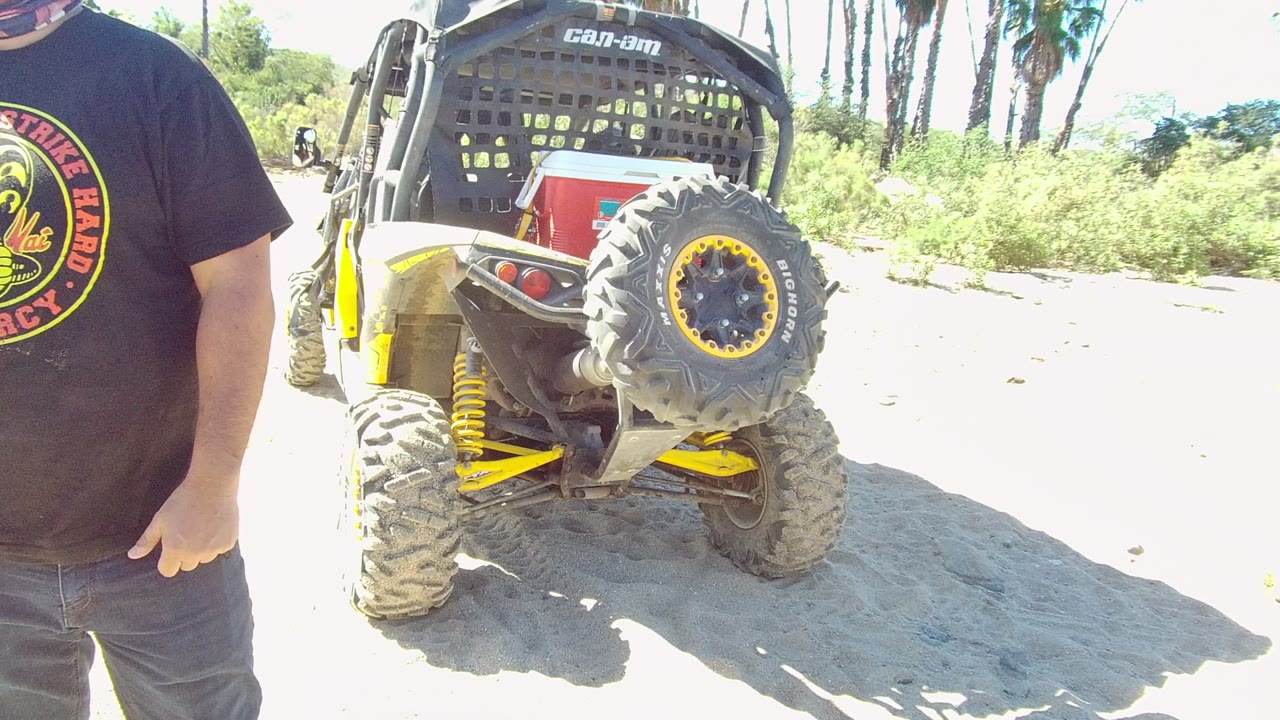 In Los Cabos, Baja California Sur, a cropped man in a black t-shirt stands partly in frame beside the rear of a yellow and black off-road vehicle on sandy ground.