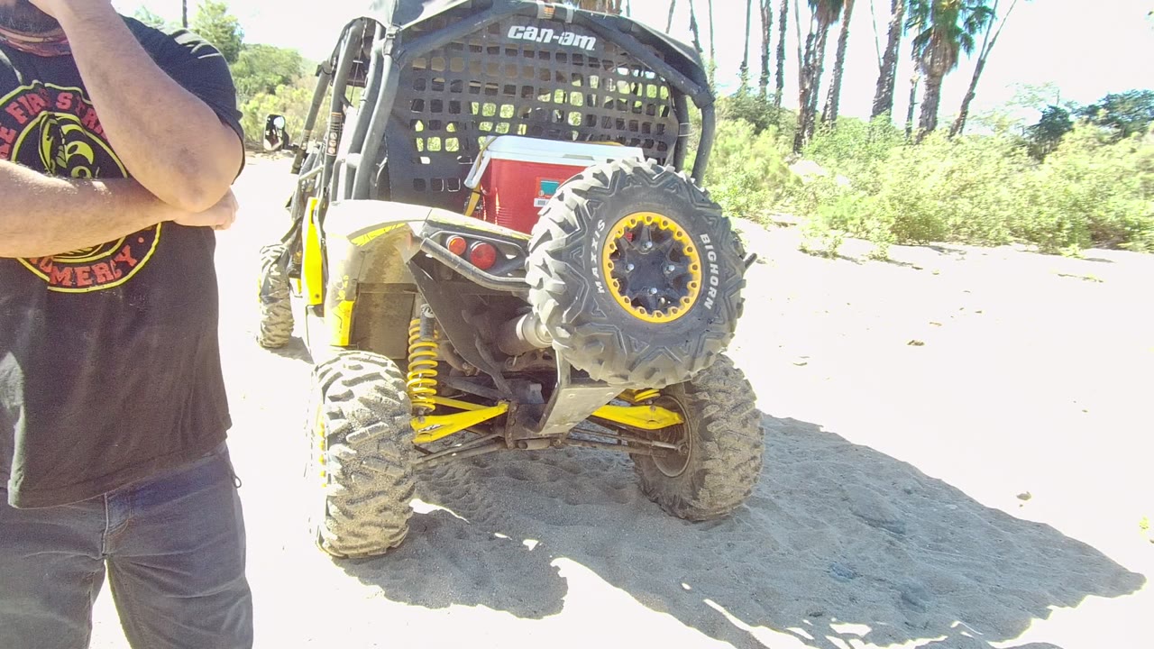 At 0:00 in Los Cabos, a cropped man in a black T-shirt stands near his face at the left edge while a Can-Am off-road vehicle sits on bright sandy ground.