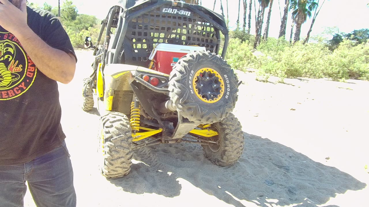At 0:02 in Los Cabos, the cropped man raises his right arm near the top of frame beside the rear of the Can-Am vehicle on overexposed sand and low greenery.
