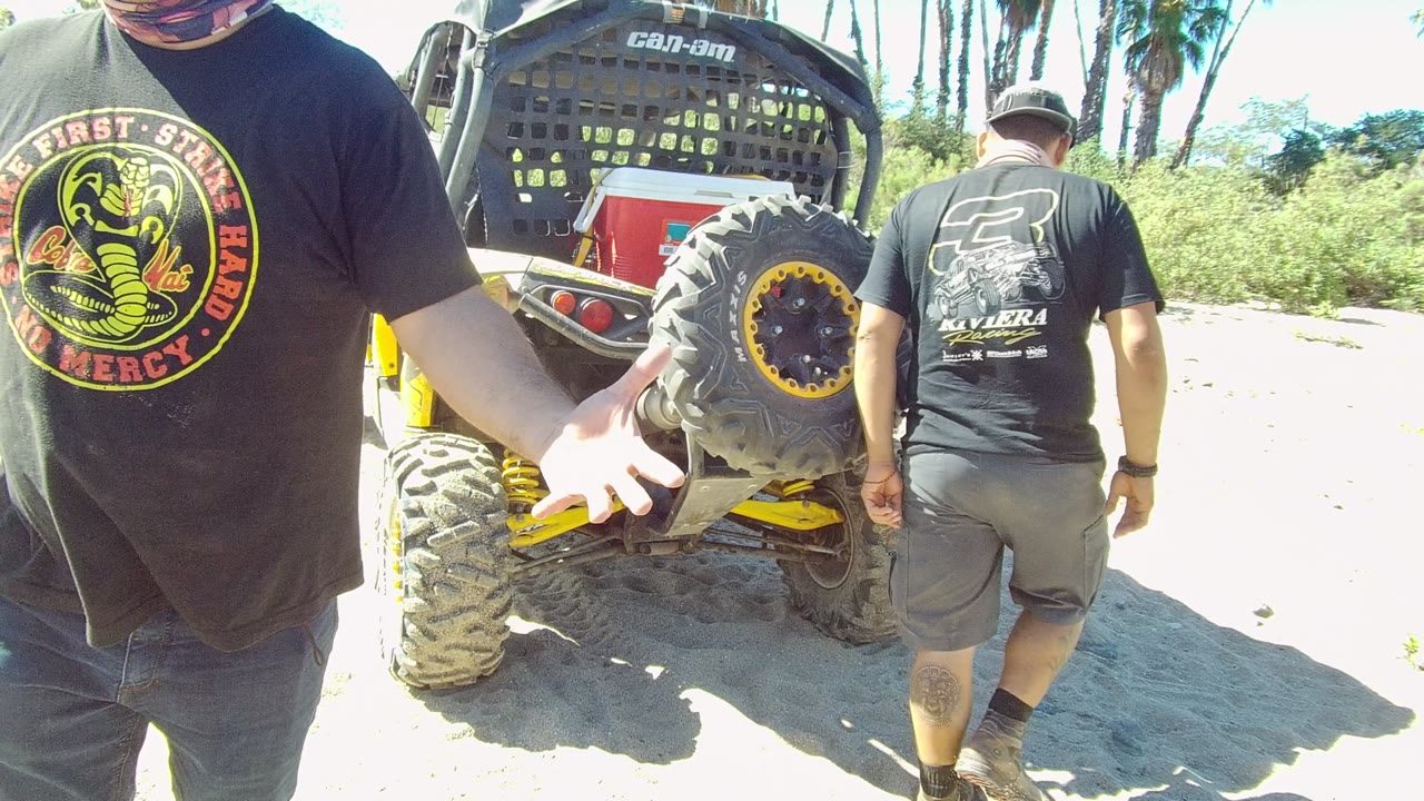 Two men stand near the rear of a yellow and black off-road vehicle on sandy ground in Los Cabos, with one cropped in the foreground and another facing away beside the tire.