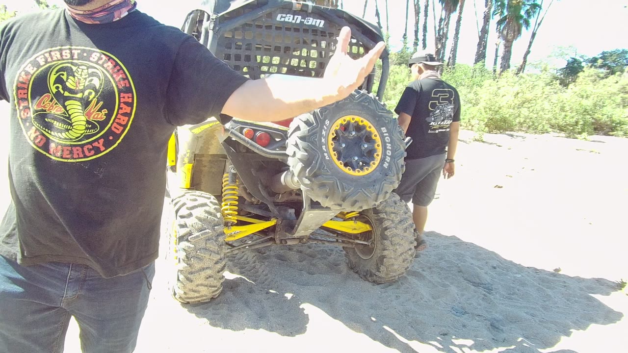 The foreground man keeps his arm raised near the rear tire of the off-road vehicle in Los Cabos, with the second man farther right and palm trunks behind the sandy track.