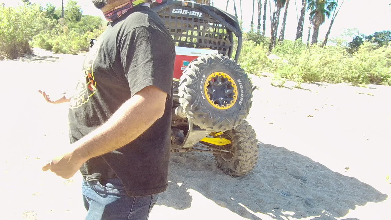 In Los Cabos, Baja California Sur, a man partly cropped at the head stands close to camera beside a yellow and black off-road vehicle with palms and greenery behind him.