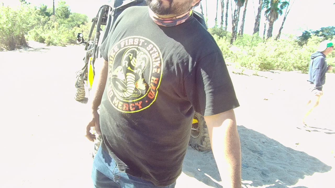 In Los Cabos, a man in a black graphic T-shirt and blue jeans stands near the camera on bright sandy ground beside a yellow-accented off-road vehicle.